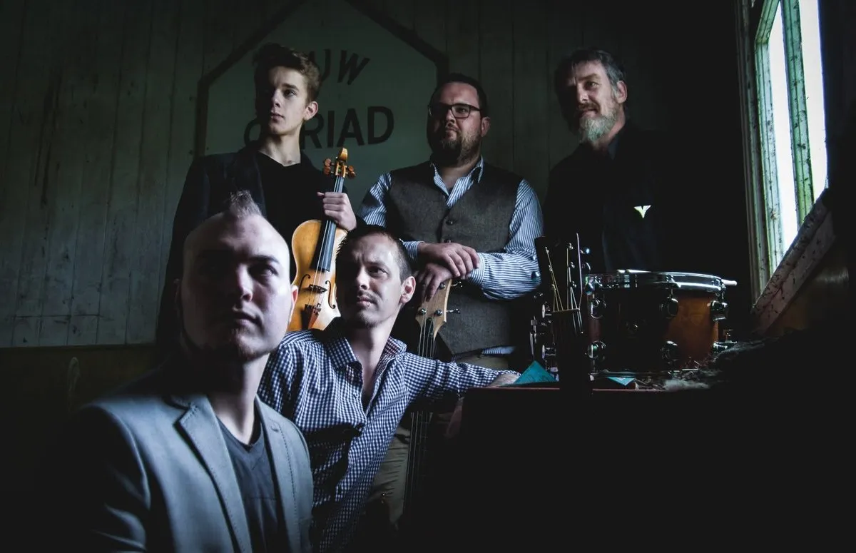 The five-piece Cats and Crows line-up of 2016 posed in a weathered wooden room, one holding a violin, a snare drum and an acoustic guitar beside them.