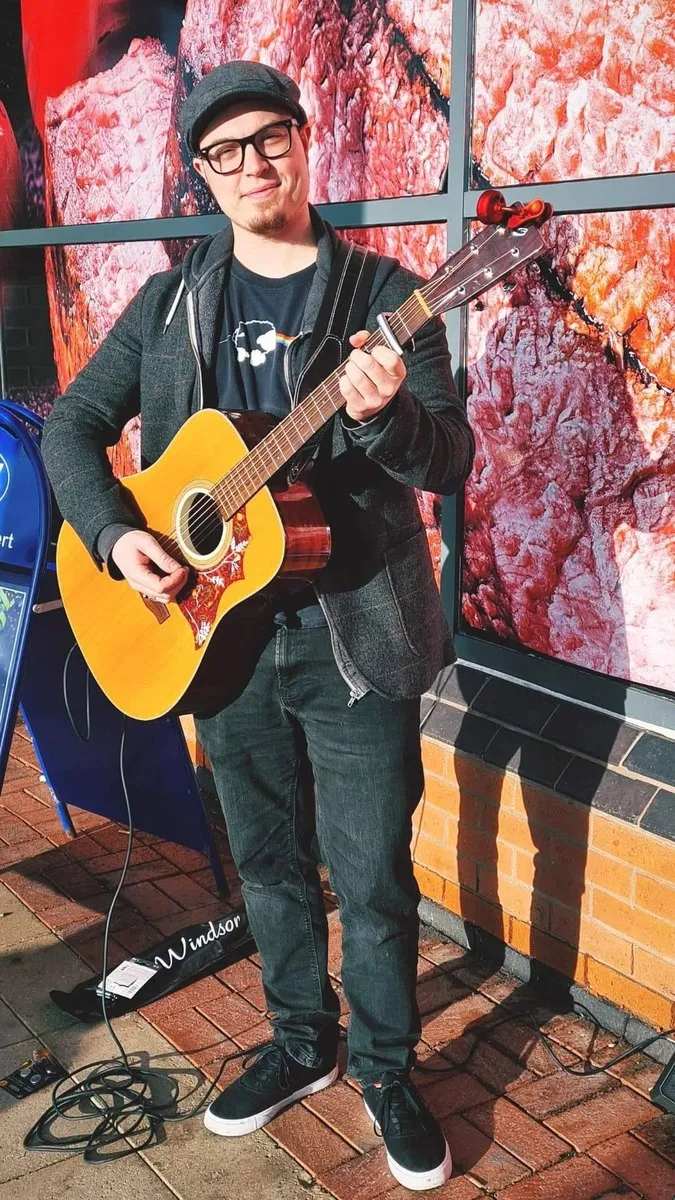 Isaac Birchall standing with an acoustic guitar outside a shop window, guitar case and cables on the pavement.