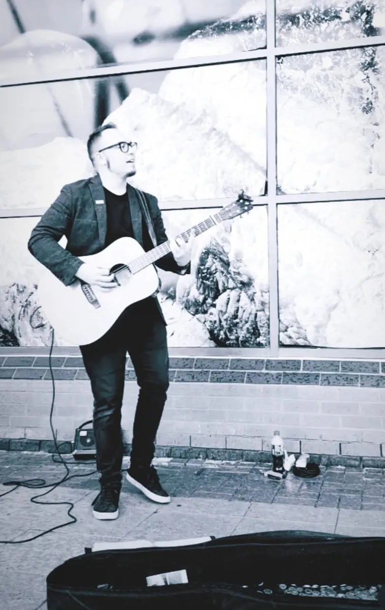 Black and white photograph of Isaac Birchall playing acoustic guitar outside a large shop window, his open guitar case on the pavement.