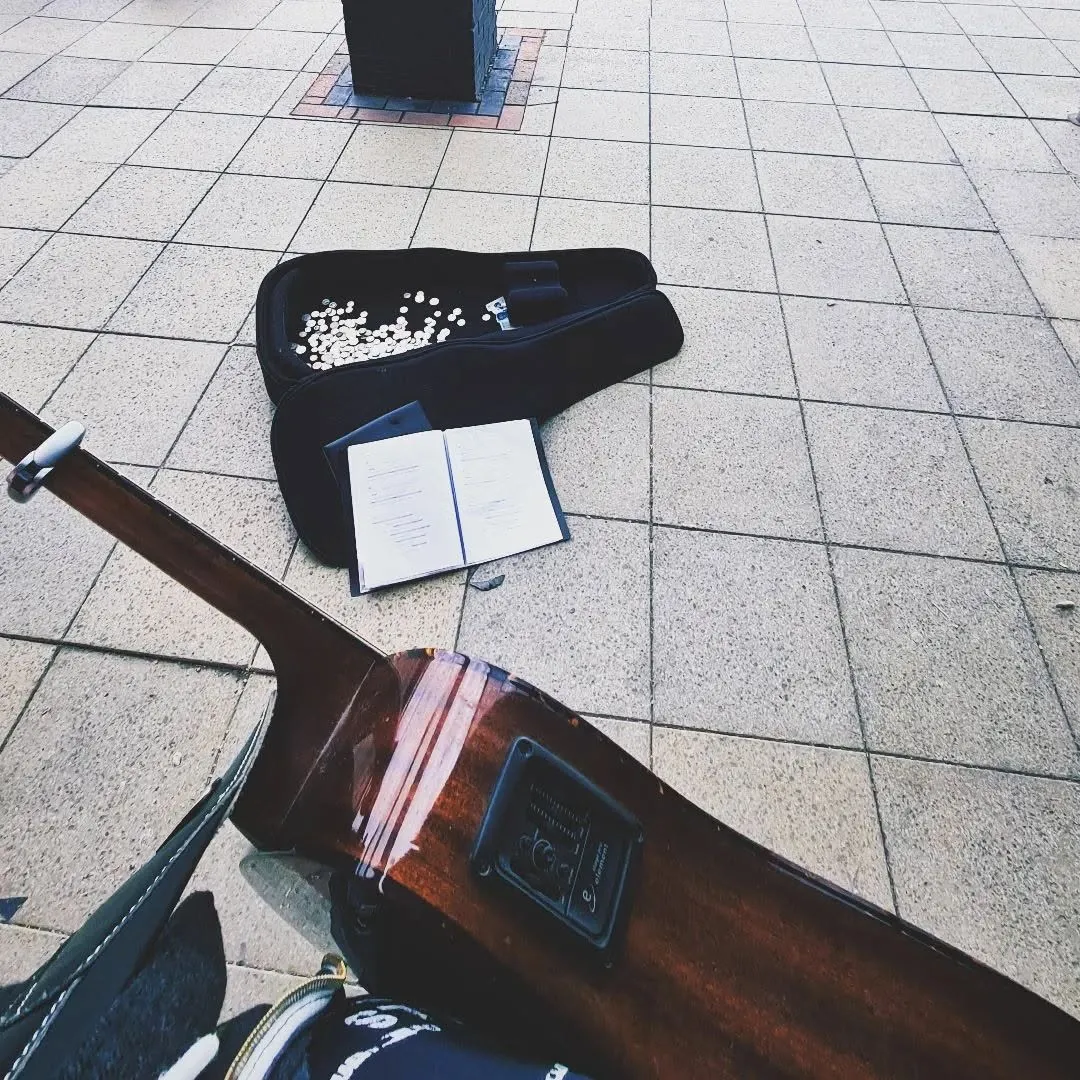 A guitar case open on paving slabs with coins in it and a notebook of songs beside it, the neck of an acoustic guitar in the foreground.