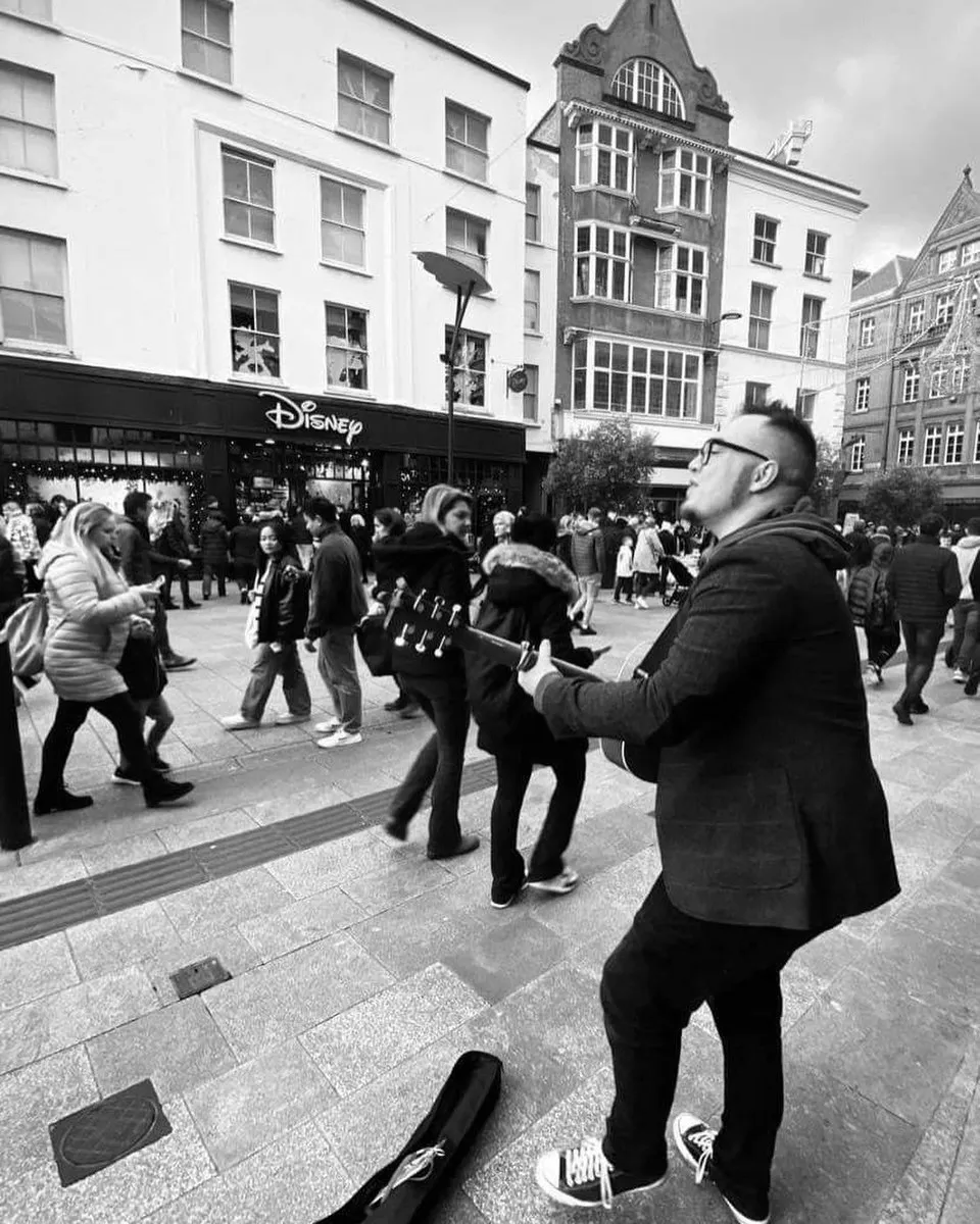 Black and white photograph of Isaac Birchall busking on a crowded Dublin street with his guitar case open in front of him, shoppers passing behind.