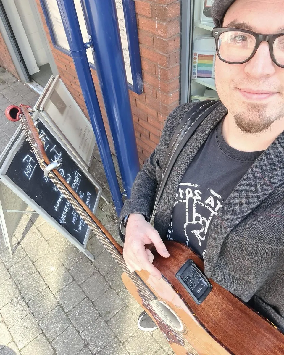 Isaac Birchall smiling at the camera with his acoustic guitar, a chalkboard sign and shop front behind him.