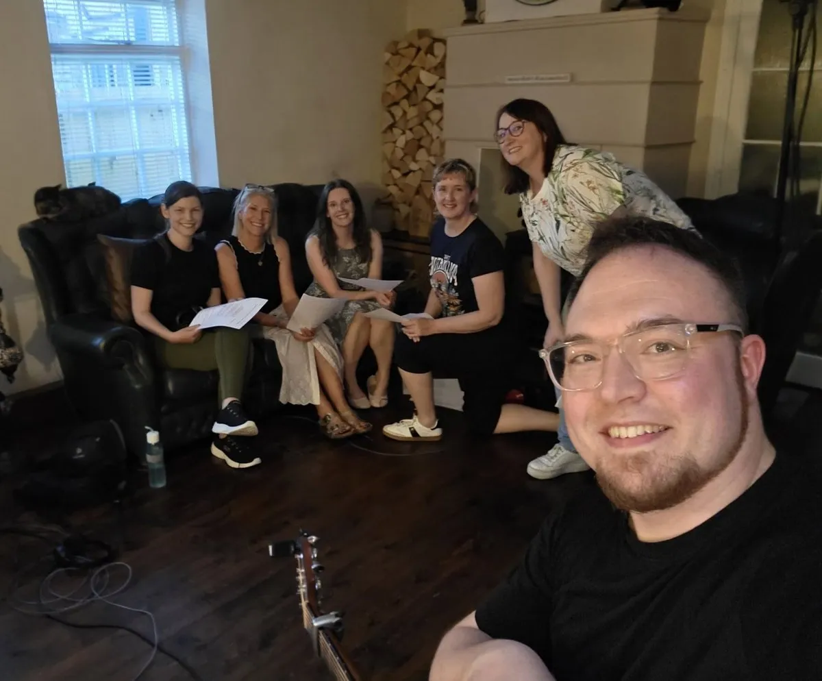 Isaac Birchall grinning at the camera while five singers sit on a sofa behind him holding lyric sheets, a guitar and a microphone stand in the foreground.