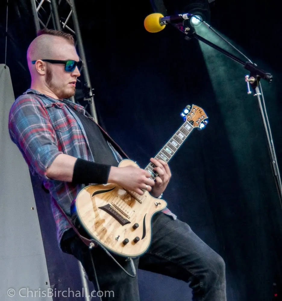 Isaac Birchall in sunglasses leaning back with a natural finish electric guitar on an outdoor festival stage.