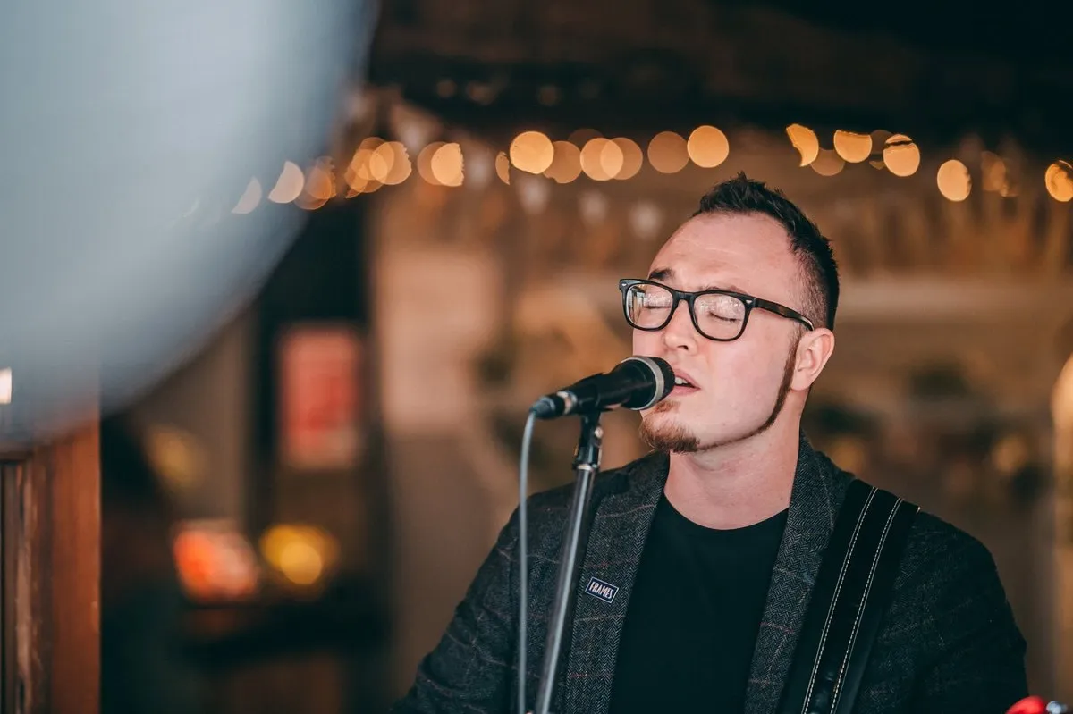 Isaac Birchall in glasses singing into a microphone, warm string lights out of focus behind him.