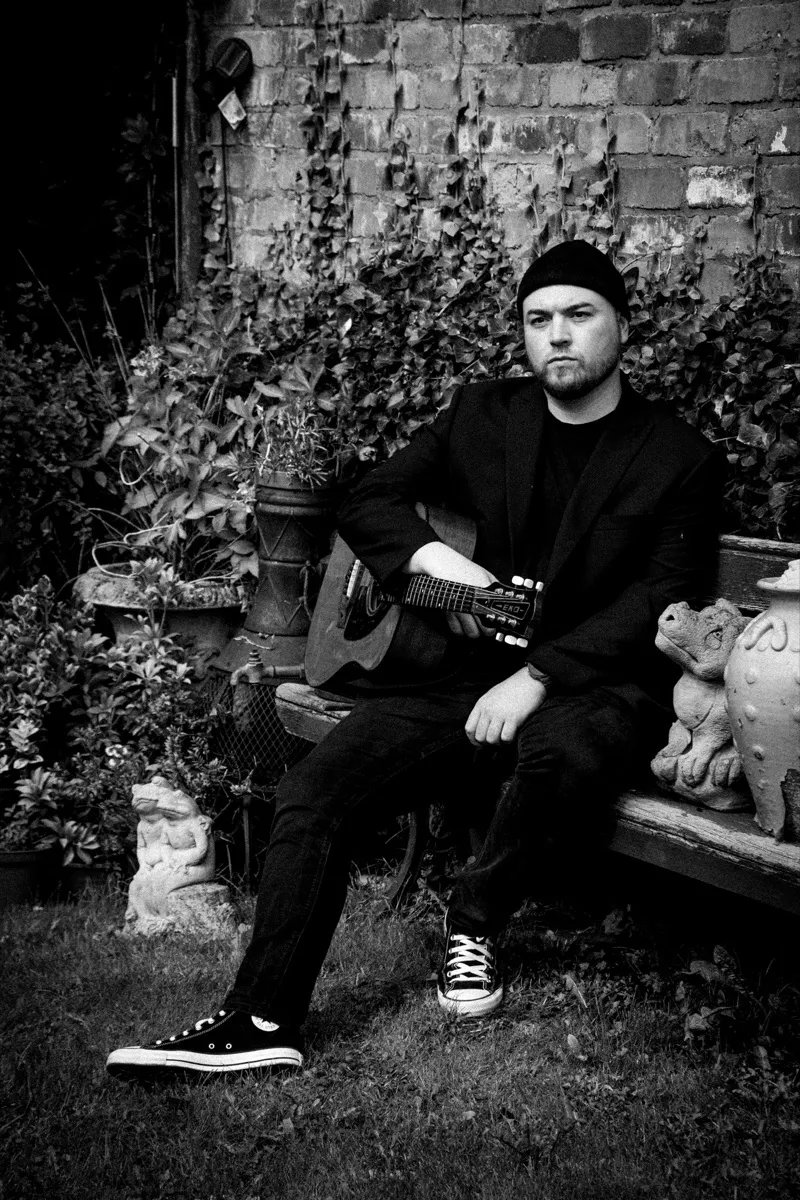 Black and white photograph of Isaac Birchall sitting on a weathered garden bench in a black jacket and Converse, an acoustic guitar across his lap and stone ornaments beside him.