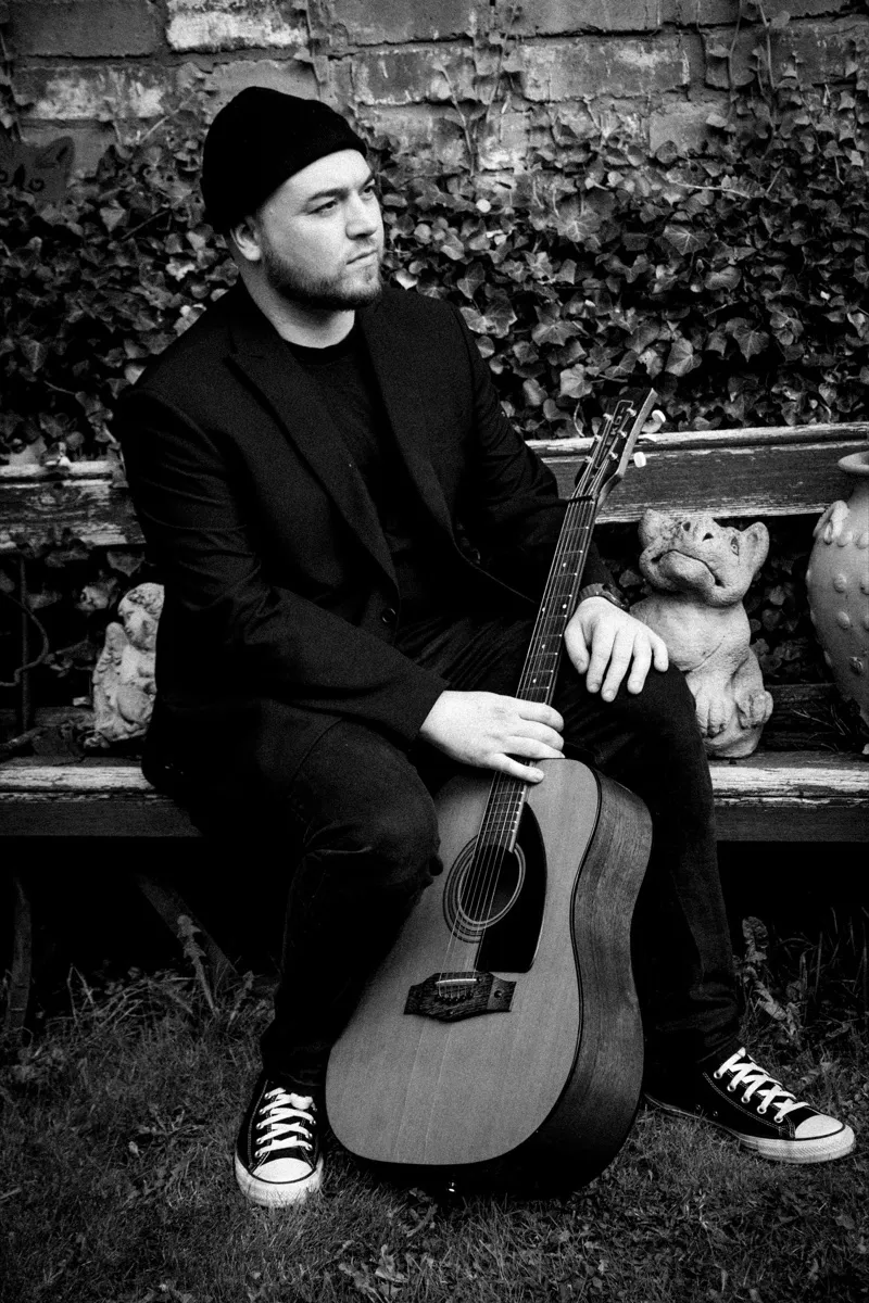 Black and white photograph of Isaac Birchall sitting on a garden bench in front of an ivy covered brick wall, his acoustic guitar standing upright between his knees.