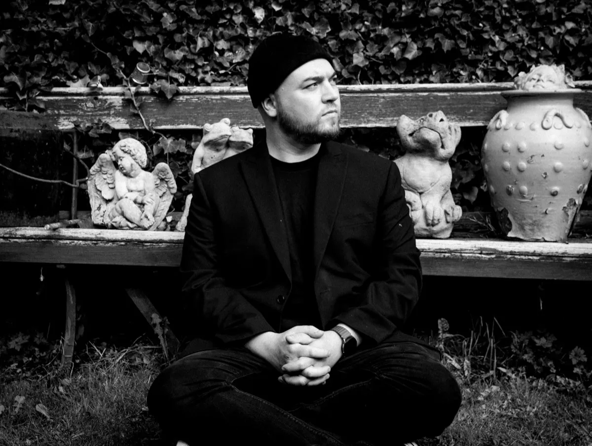 Black and white photograph of Isaac Birchall sitting cross legged on the grass in front of a weathered bench lined with a stone cherub, a frog and a pot, ivy on the wall behind.