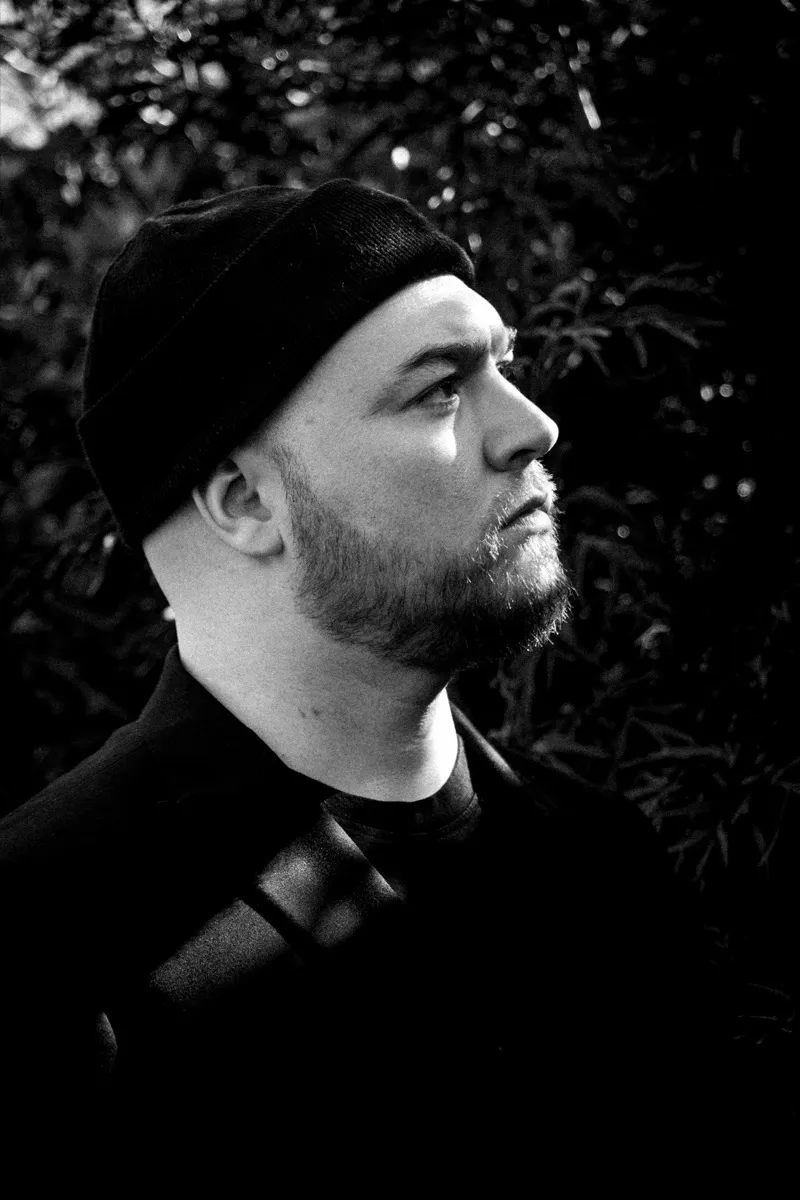 Black and white portrait of Isaac Birchall in a black beanie, chin lifted and looking up, out of focus leaves behind him.