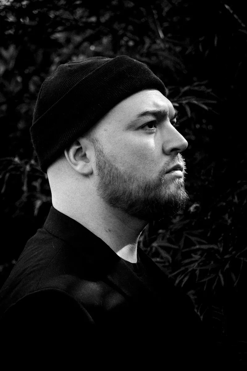 Black and white close profile of Isaac Birchall in a black beanie and jacket, looking off to one side against dark garden foliage.