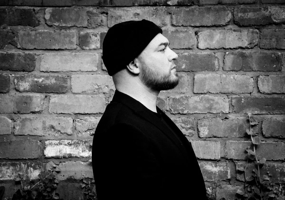 Black and white profile of Isaac Birchall in a black beanie and jacket standing against an old brick wall with ivy creeping up it.