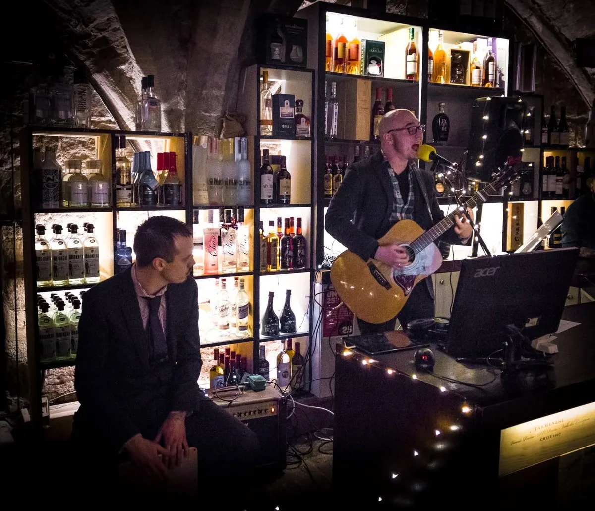 Isaac Birchall singing with an acoustic guitar in a bar lined with lit shelves of bottles, Andy Cooper sitting on a cajon beside him.