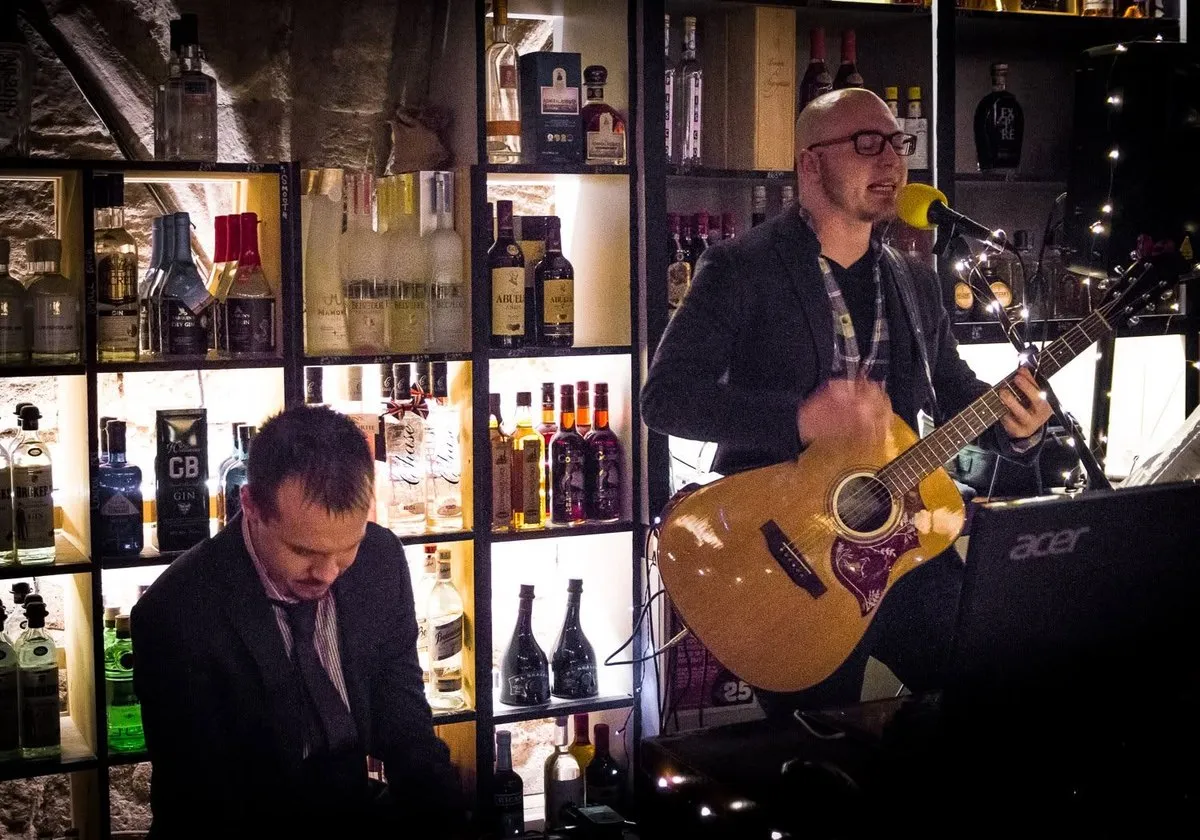 Andy Cooper playing cajon in the foreground while Isaac Birchall sings with an acoustic guitar, lit shelves of bottles behind them.