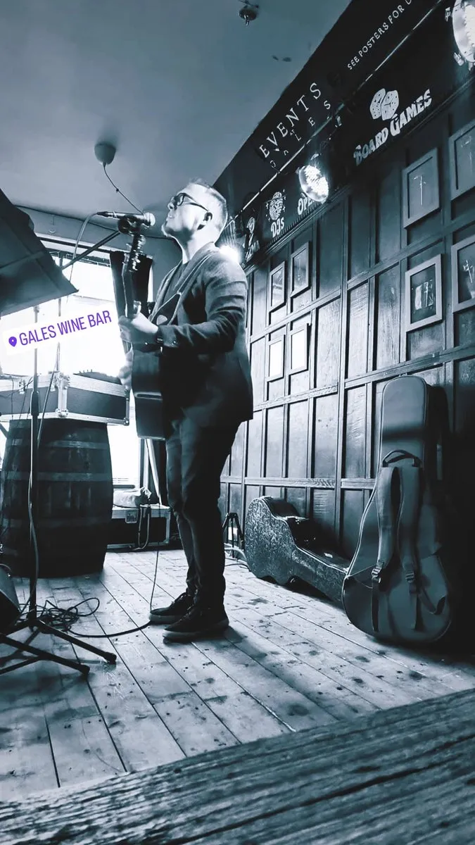 Black and white photograph of Isaac Birchall singing with an acoustic guitar in a wood panelled bar, a guitar case and barrel beside him.