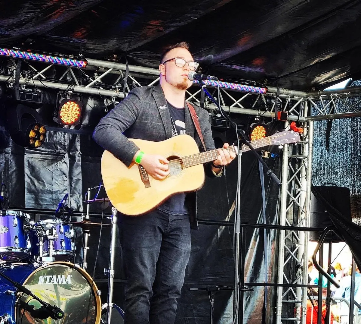 Isaac Birchall singing into a microphone with an acoustic guitar on a festival stage, lighting rig overhead and a drum kit behind him.