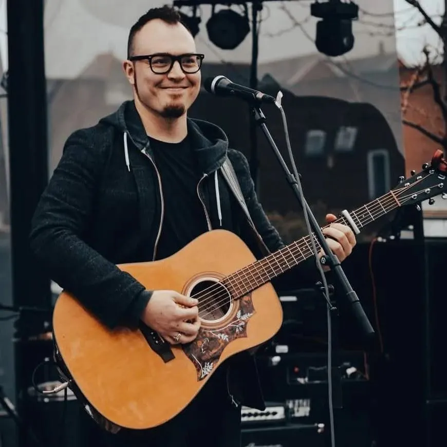 Isaac Birchall smiling with his acoustic guitar at a microphone on an outdoor stage.