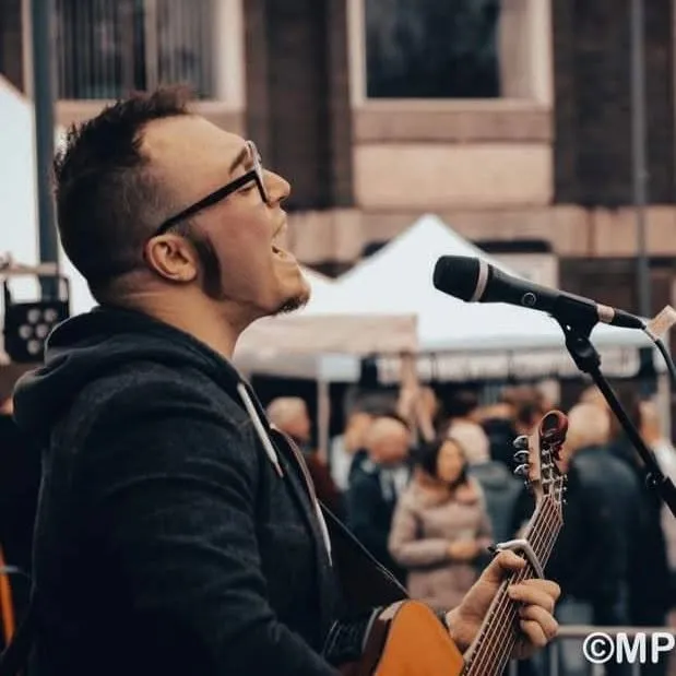 Isaac Birchall singing into a microphone with an acoustic guitar at an outdoor market, a crowd and stalls behind him.