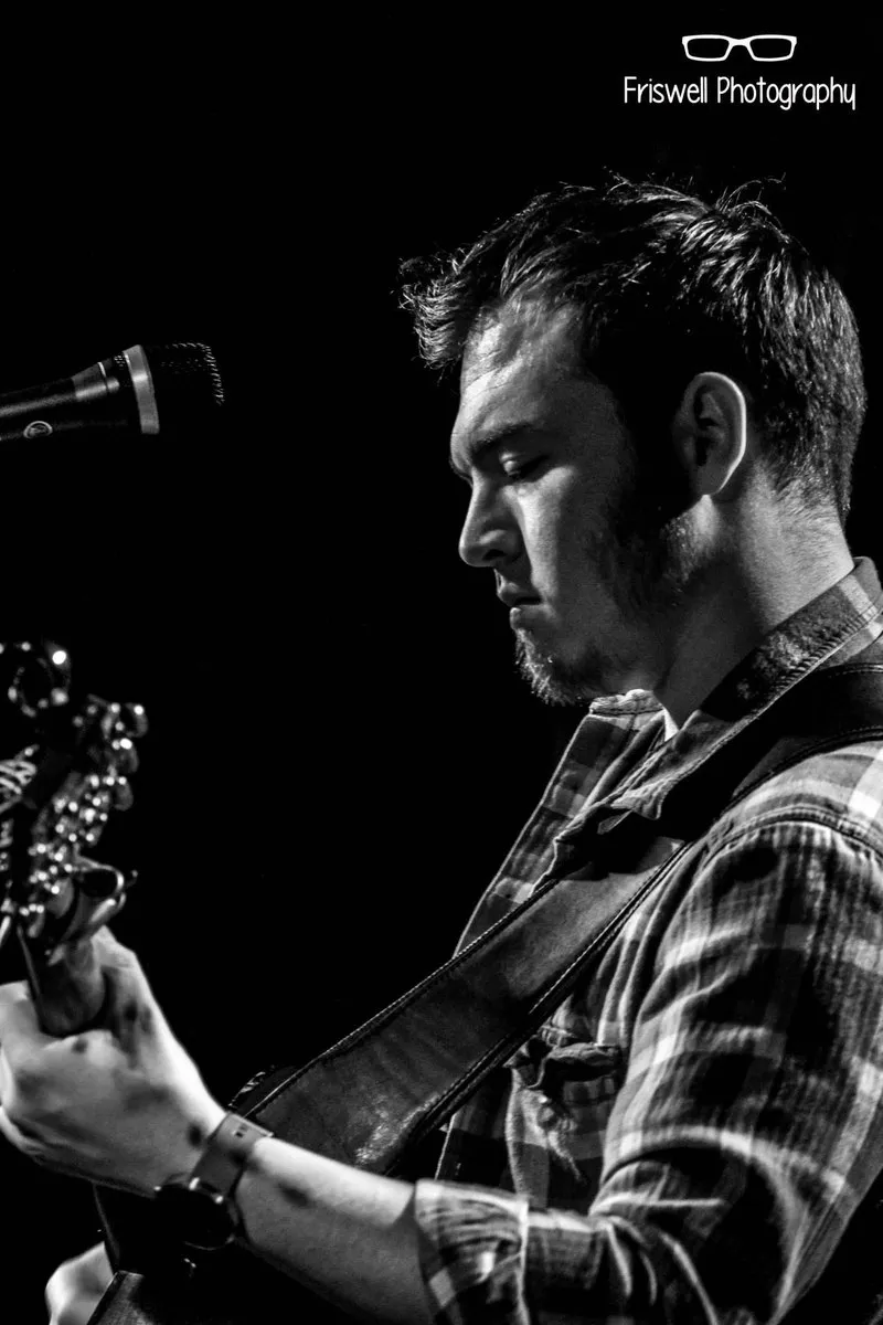 Black and white profile of Isaac Birchall at a microphone with his guitar, lit against a dark background.