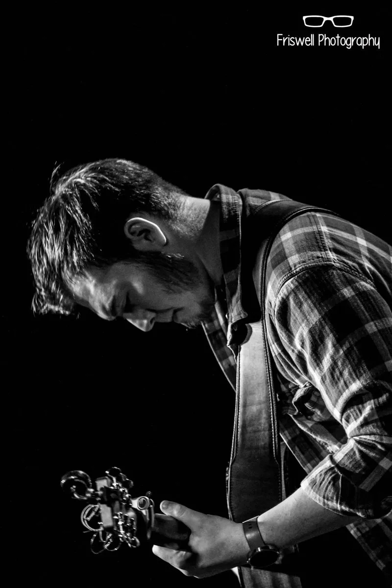 Black and white photograph of Isaac Birchall looking down over the neck of his guitar against a dark background.