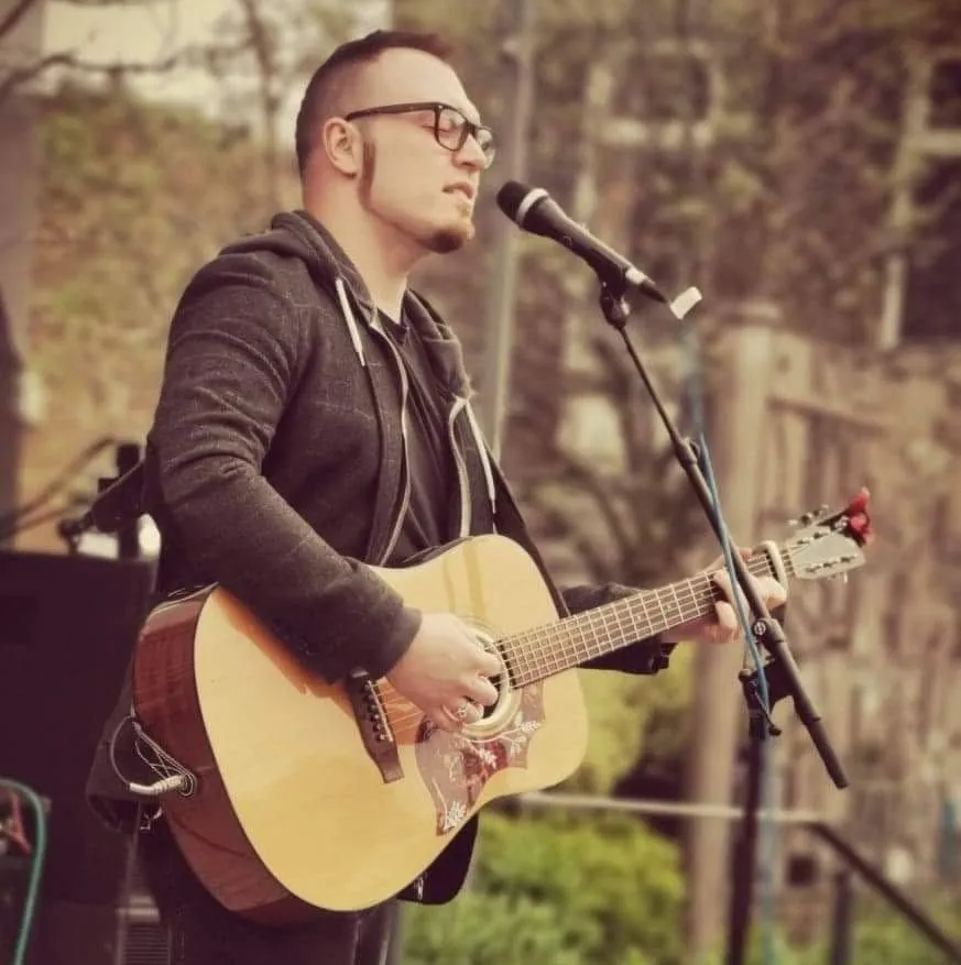 Isaac Birchall singing into a microphone with an acoustic guitar on an outdoor stage, trees behind him.