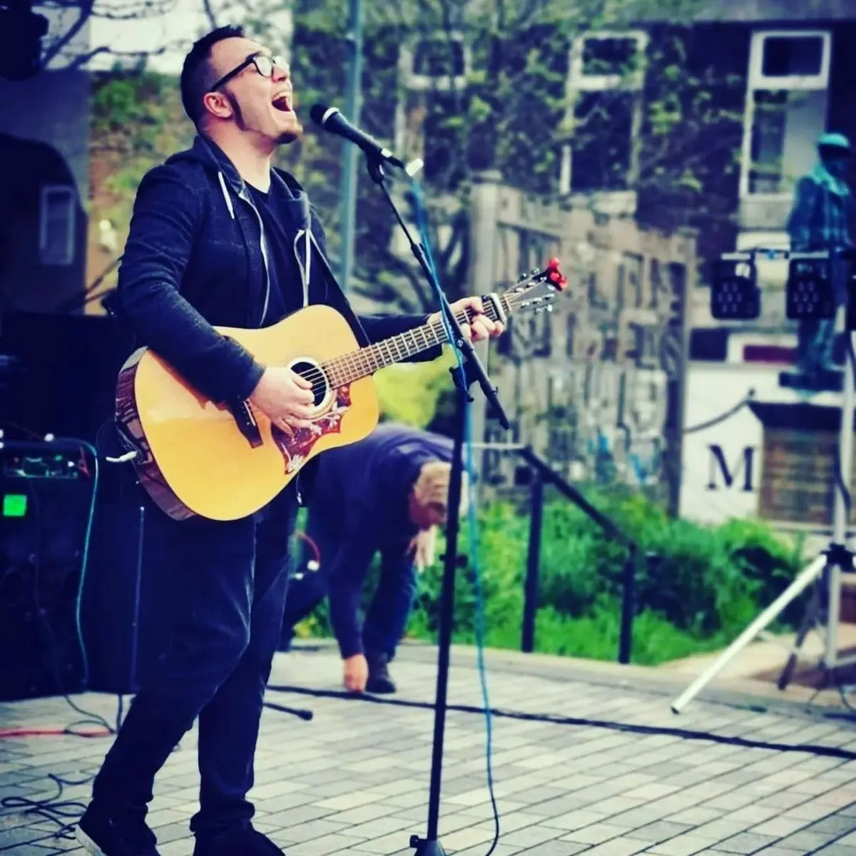 Isaac Birchall singing with his head back at an outdoor microphone, an acoustic guitar in his hands and a statue behind him.