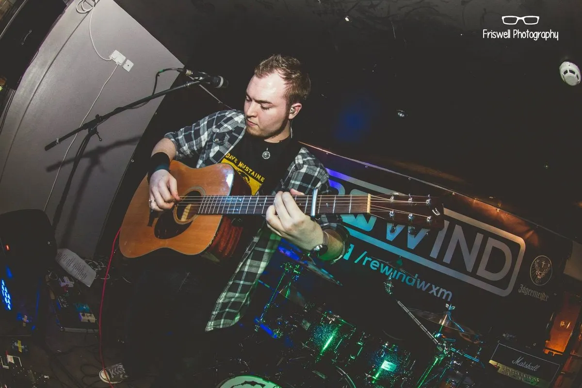 Wide angle photograph looking down on Isaac Birchall playing acoustic guitar in front of a drum kit, a Rewind backdrop behind him.