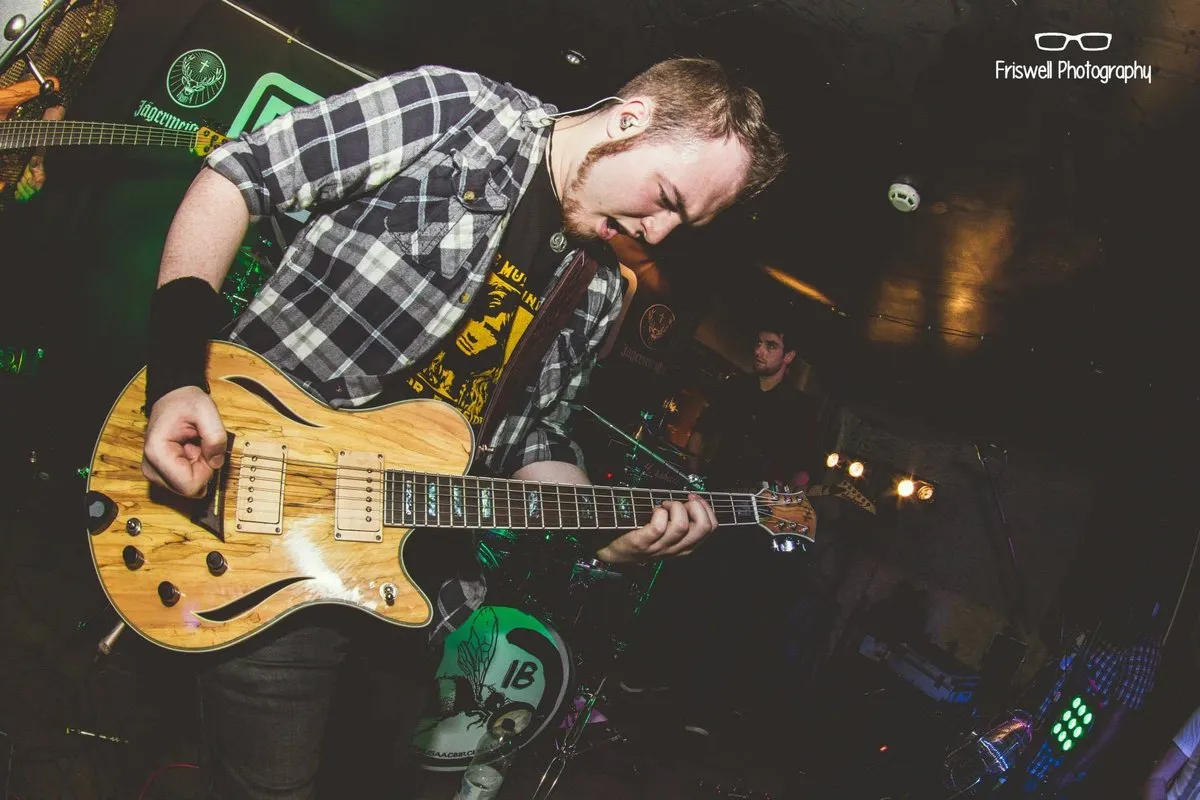 Isaac Birchall bent over his guitar mid song under green stage light, a drummer playing behind him.