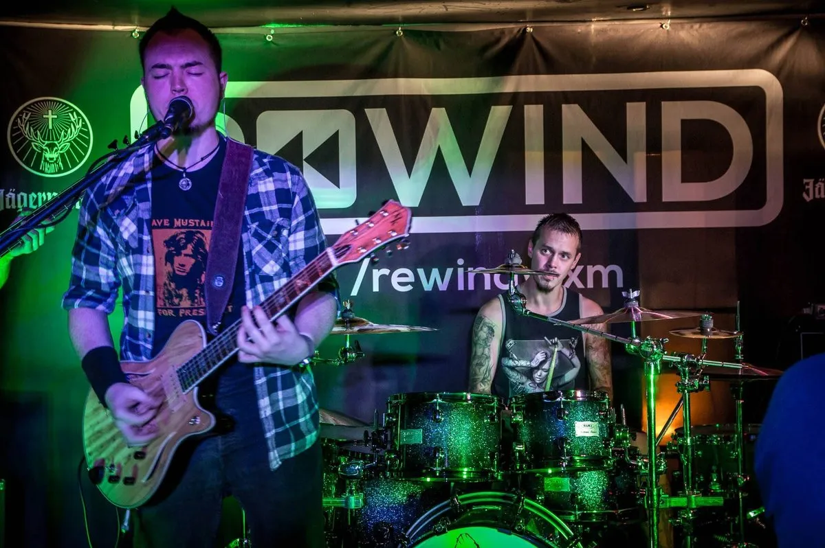 Isaac Birchall singing and playing electric guitar under green stage lights, a drummer behind him and a Rewind backdrop on the wall.