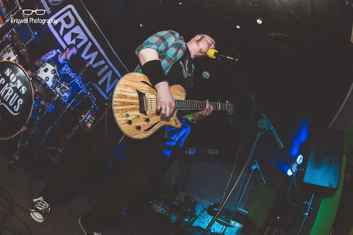 Isaac Birchall leaning over his guitar at the microphone, a drummer behind him and a Rewind backdrop on the wall.