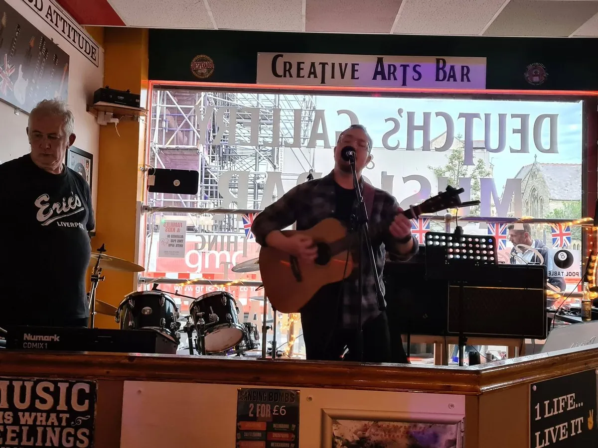 Isaac Birchall singing and playing acoustic guitar behind a bar, a Creative Arts Bar sign above him and a drum kit to one side.