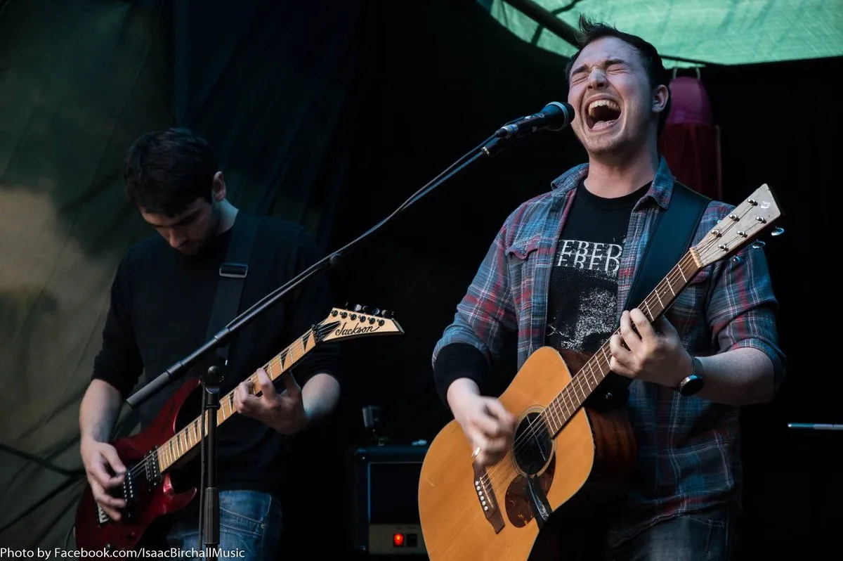 Isaac Birchall singing hard into a microphone with his eyes shut, playing acoustic guitar beside another guitarist on an outdoor stage.