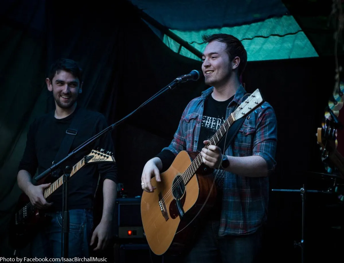Isaac Birchall and another guitarist smiling on an outdoor stage under a green tarpaulin, Isaac at the microphone with his acoustic guitar.