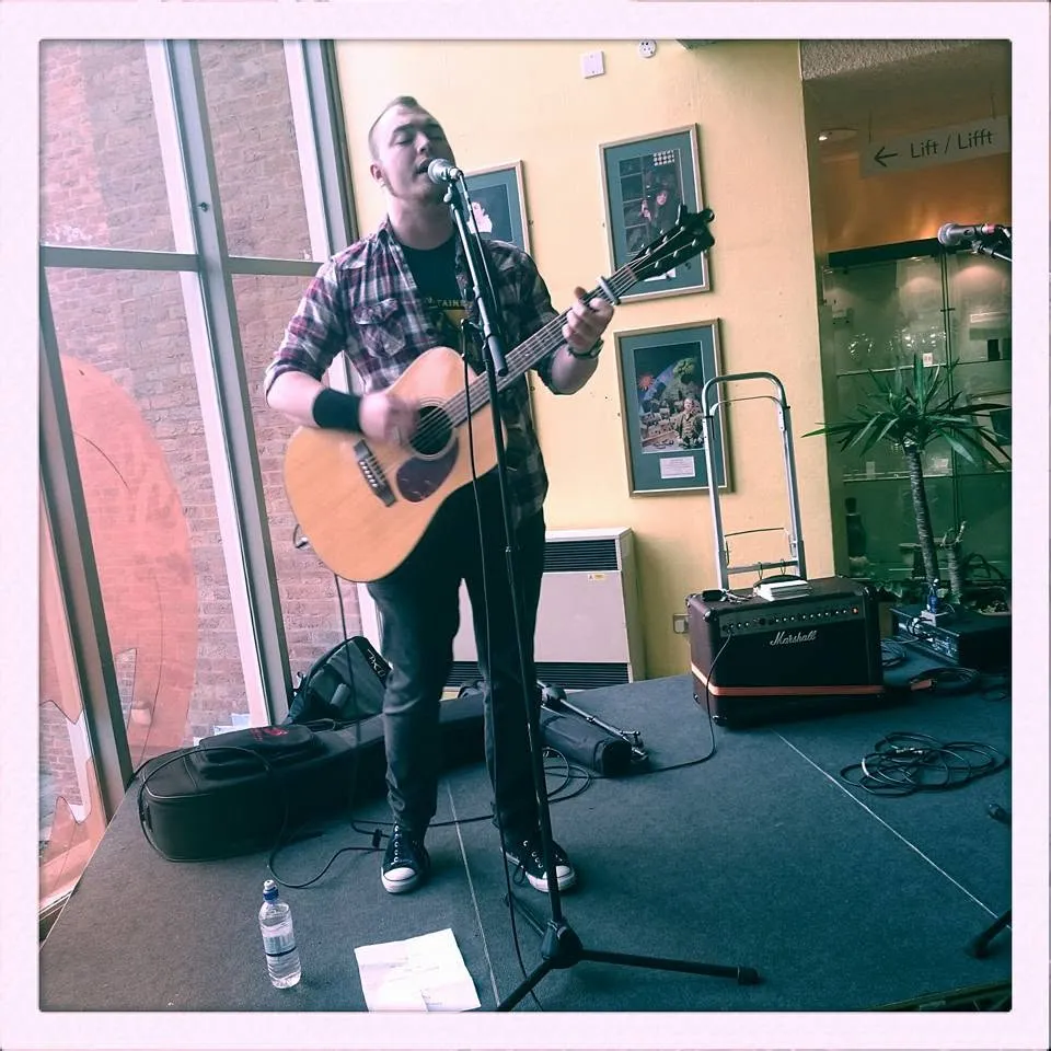 Isaac Birchall alone on a small indoor stage with an acoustic guitar, an amplifier behind him and a bilingual lift sign on the wall.